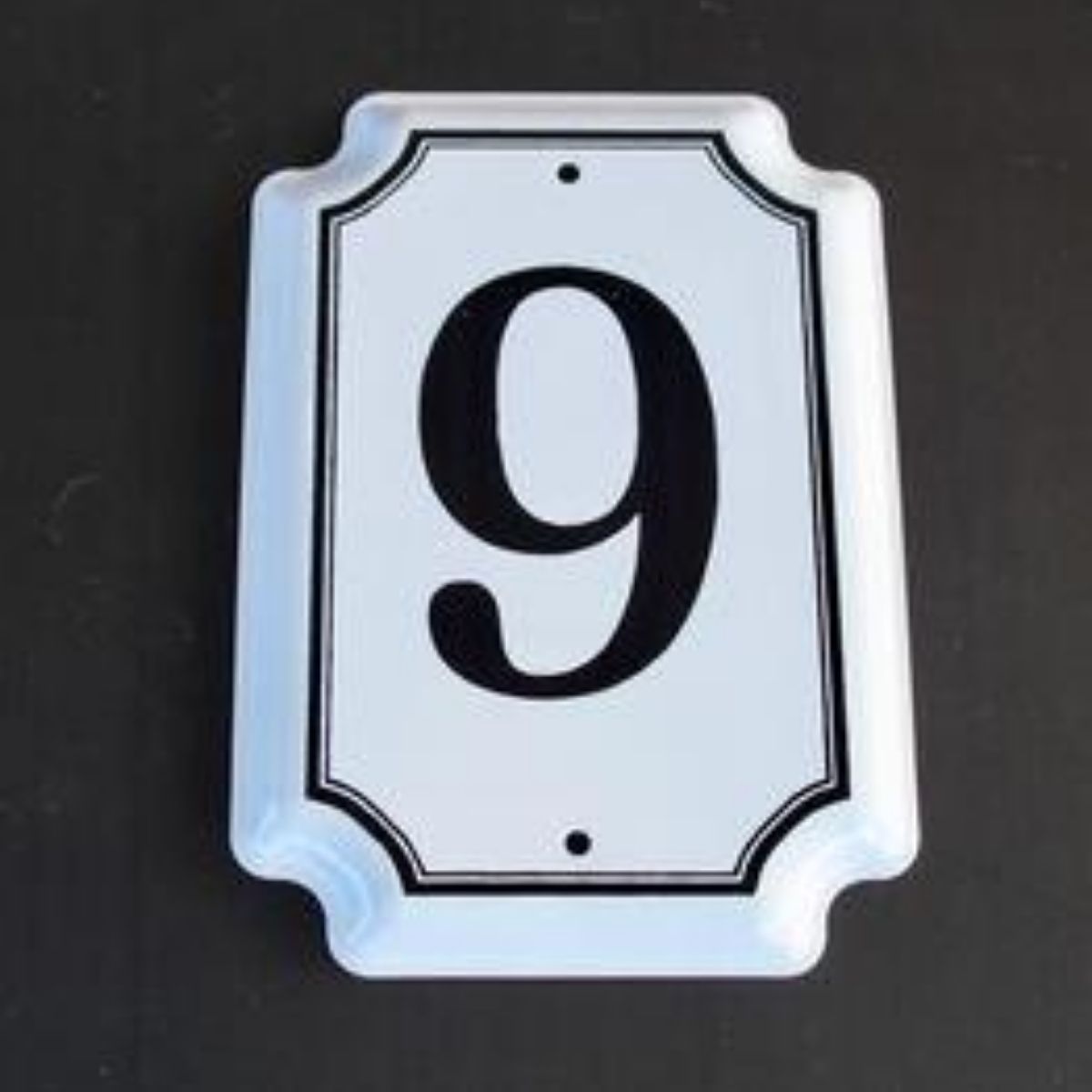 Traditional white enamel house number with the numeral  in black, with a  white border and screw holes visible.